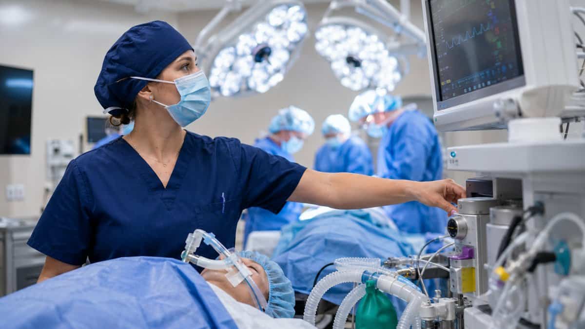 Certified registered nurse anesthetist at the head of the operating table adjusting the anesthesia machine while watching the patient monitor during surgery